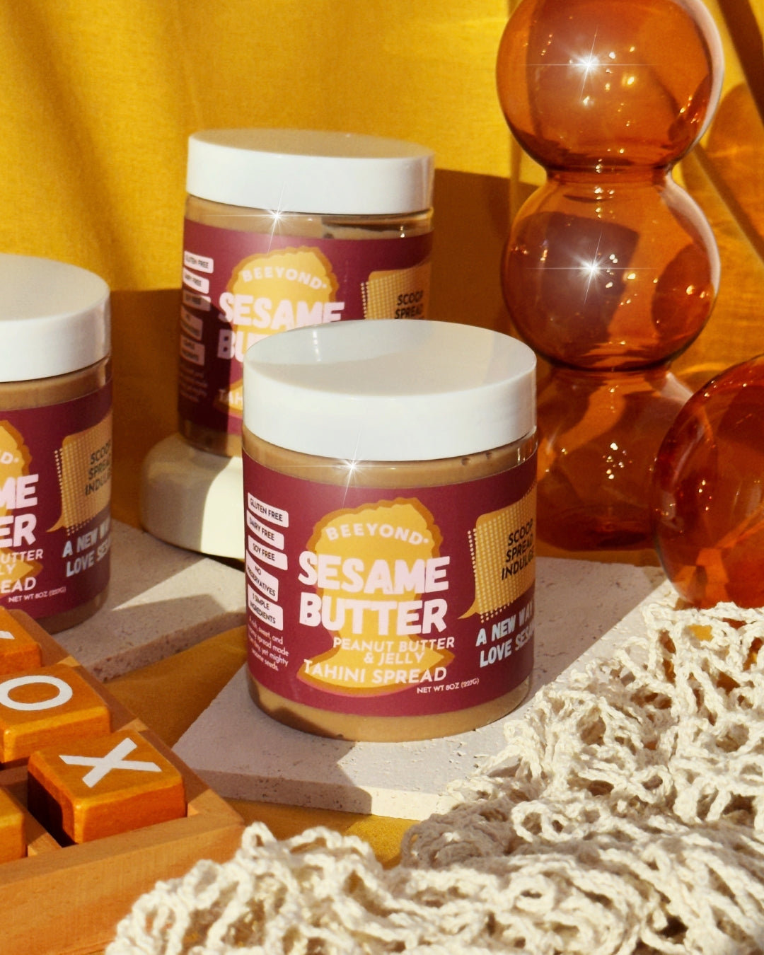 Three jars of Peanut Buttter & Jelly sesame butter sitting on stone slabs and surrounded by various objects just as a netted produce bag, a cylindrical orange vase, and a tic tac toe board