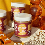 Three jars of Peanut Buttter & Jelly sesame butter sitting on stone slabs and surrounded by various objects just as a netted produce bag, a cylindrical orange vase, and a tic tac toe board